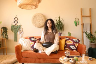 Genç Afro-Amerikan cadı glukofon ile evdeki kanepede meditasyon yapıyor.
