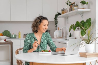 Bilgisayarlı olgun bir kadın mutfakta sebze salatası yiyor.