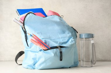 Masada beyaz grunge duvarına karşı kırtasiye malzemesi ve bir şişe su olan mavi sırt çantası.