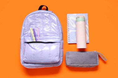 Okul malzemeleriyle birlikte leylak rengi sırt çantası ve turuncu arka planda bir şişe su. Üst görünüm