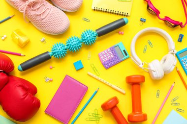 Masaj çubuğu, kulaklıklar, boks eldivenleri, halterler, spor ayakkabıları ve sarı arka planda farklı kırtasiye malzemeleri.