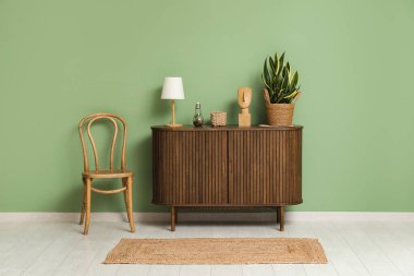 Koyu ahşap çekmeceler, yeşil duvarın yanında lamba ve çiçek var.