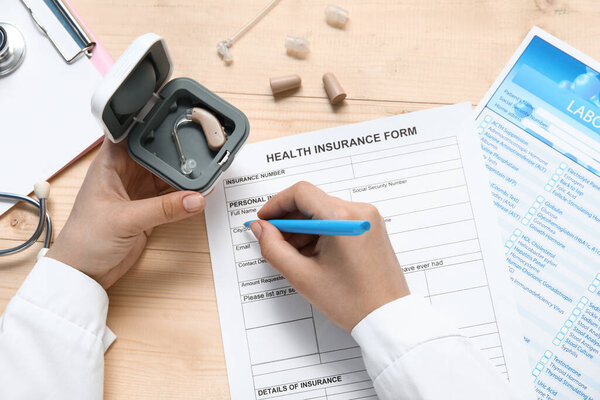 Female doctor with hearing aid writing in health insurance form on wooden background, closeup
