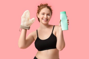 Güzel, genç ve mutlu bir kadın, banyo masaj eldiveni ve pembe arka planda bir şişe duş jeli.