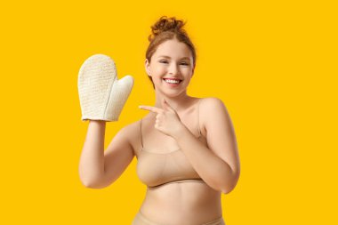 Güzel, genç ve mutlu bir kadın sarı arka planda banyo masajı eldivenini işaret ediyor.