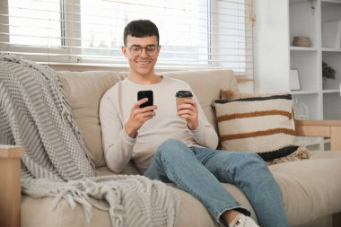 Moda gözlüklü, cep telefonlu ve evdeki kanepede oturan yakışıklı, mutlu genç adam.