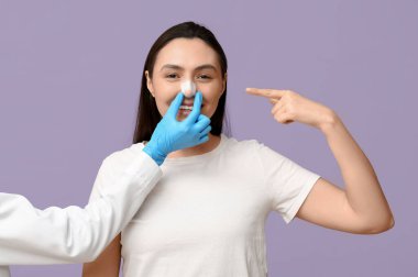 Estetik cerrah eli ve genç bir kadın leylak arka planındaki sargılı burnu işaret ediyor.