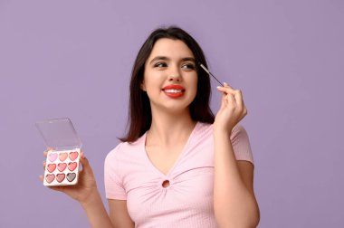 Ruj paleti ve leylak arkaplanda makyaj fırçası olan güzel bir genç kadın.