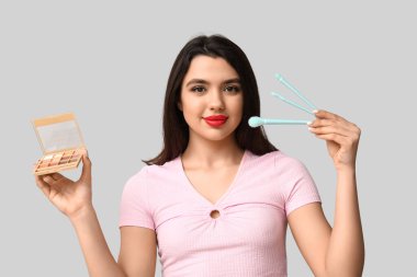 Göz falı paleti ve gri arka planda makyaj fırçaları olan güzel bir genç kadın.