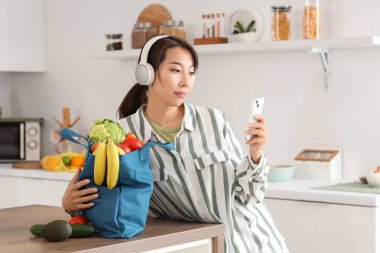 Kulaklıklı genç Asyalı kadın mutfaktaki alışveriş çantasının yanında cep telefonu kullanıyor.