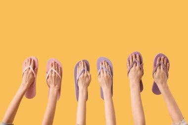 Sarı arka planda üç çift parmak arası terlik tutan eller.