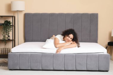 Yatak odasındaki yumuşak yatağın üzerinde yastıklı mutlu Afro-Amerikan kadın.