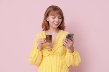 Bir bardak lezzetli buzlu latte ve pembe arka planda cep telefonu olan mutlu genç bir kadın.