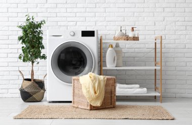 Beyaz tuğla duvarın yanında rafı, evi ve sepeti olan modern çamaşır makinesi.