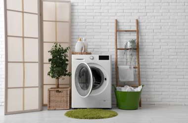 Beyaz tuğla duvarın yanında merdiveni, sepeti ve bitkisi olan modern çamaşır makinesi