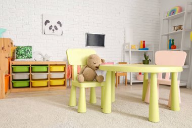 Anaokulundaki oyun odasının masasının yanında oyuncak ayı ve rafların yanında oturuyordu.