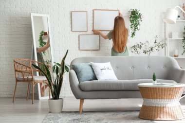 Genç bir kadın oturma odasındaki beyaz tuğla duvara boş çerçeveler asıyor.