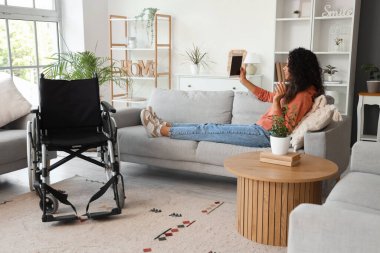 Tekerlekli sandalyesi ve tablet bilgisayarı olan genç bir Afro-Amerikalı kadın evdeki kanepede yatıyor.