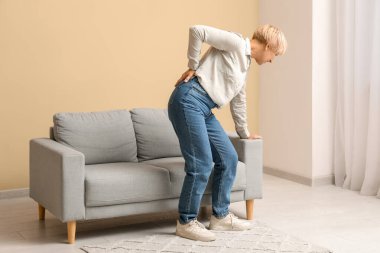 Oturma odasında kanepeden kalkarken sırt ağrısı çeken olgun bir kadın.