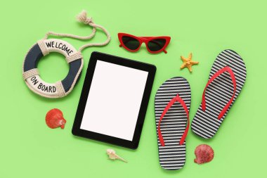 Dekor cankurtaran şamandıralı tablet ve plaj aksesuarları ve yeşil arka planda parmak arası terlikler. Üst görünüm