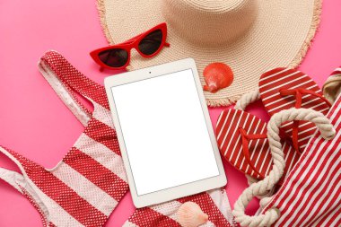 Tablet ve plaj aksesuarları, pembe arka planda mayo ve parmak arası terlikler. Üst görünüm