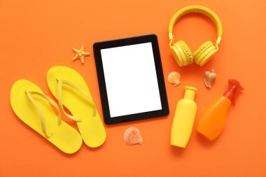 Tablet ve plaj aksesuarları, parmak arası terlikler ve turuncu arka planda kulaklıklar. Üst görünüm