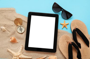 Tablet ve plaj aksesuarları, parmak arası terlik, güneş gözlüğü ve mavi arka planda kum. Üst görünüm