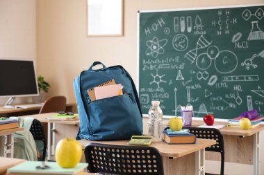 Kitaplı elma, su şişesi ve sırt çantası sınıfın masasında.