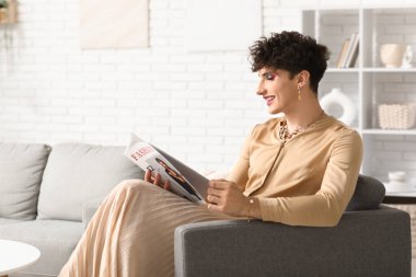 Genç transseksüel kadın evdeki koltukta dergi okuyor.