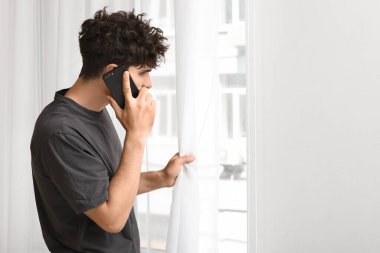 Genç adam cep telefonuyla konuşuyor. Evinin penceresinin yanındaki ışık duvarında küf var.