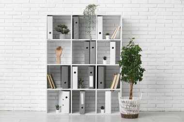 Ofiste beyaz tuğla duvarın yanında doküman klasörleri ve bitkilerle raf ünitesi