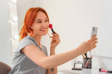 Cep telefonu ve makyaj fırçasıyla evde selfie çeken genç bir kadın.