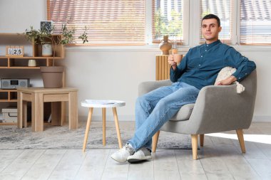 Modern oturma odasındaki koltukta oturan bir fincan kahveli yakışıklı genç adam.