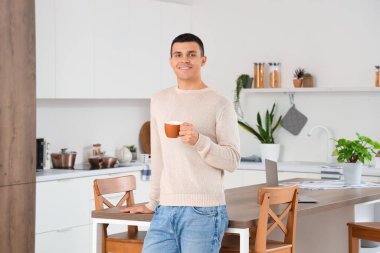 Masasında modern dizüstü bilgisayarı olan mutfakta kahve içen yakışıklı bir genç.