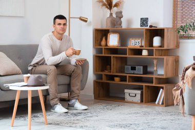 Modern oturma odasındaki kanepede oturan bir fincan kahveli yakışıklı genç adam.