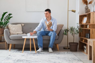 Oturma odasındaki kanepede oturan kitap ve kahveyle yakışıklı genç adam.