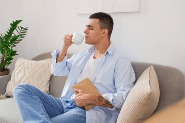 Kitaplı yakışıklı genç adam oturma odasında kanepede otururken kahve içiyor.