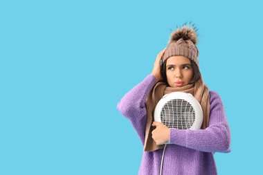Beautiful young thoughtful woman in winter clothes with electric fan heater on blue background clipart