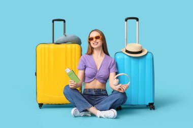 Mavi arka planda pasaportu ve kulaklığıyla bavulunun yanında oturan gülümseyen genç kadın. Seyahat kavramı