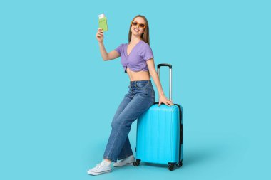 Çantanın üzerinde oturan ve elinde pasaportu olan gülümseyen genç bir kadın. Seyahat kavramı