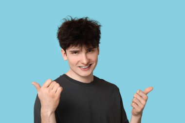 Dişlerinde diş teli olan gülümseyen genç adam mavi arka planda baş parmağını gösteriyor.
