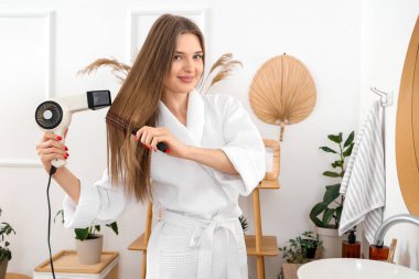 Güzel bir kadın banyoda saçlarını yuvarlak fırçayla kurutuyor.