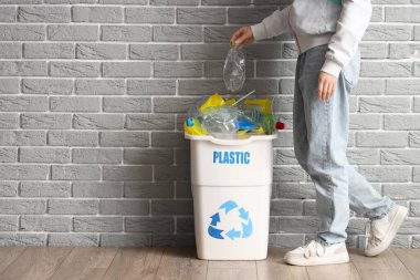 Kadın, gri tuğla duvarın yanında plastikçi işareti olan bir şişeyi çöp kutusuna koyuyor. Geri dönüşüm kavramı