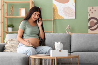 Evdeki kanepede cep telefonuyla konuşan defterli hamile bir kadın. Annelik Faydası konsepti