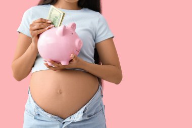 Genç hamile bir kadın pembe arka planda kumbaraya para yatırıyor, yakın plan. Annelik Faydası konsepti
