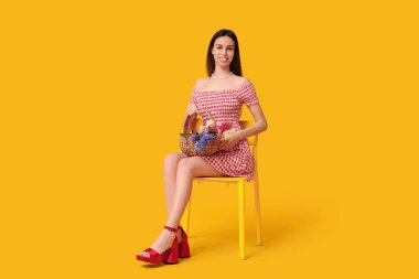 Mutlu, gülümseyen, sepetli, sümbül çiçekli genç kadın sarı arka planda, sandalyede oturuyor.