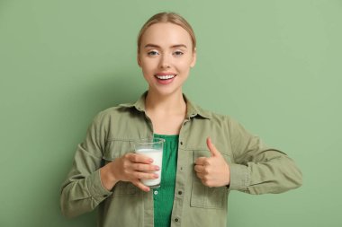 Yeşil arka planda baş parmağını gösteren bir bardak sütle güzel bir genç kadın.
