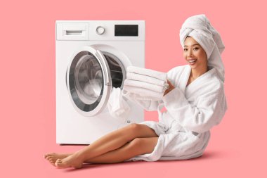 Bornozlu, temiz elbiseli güzel bir kadın pembe arka planda çamaşır makinesinin yanında oturuyor.