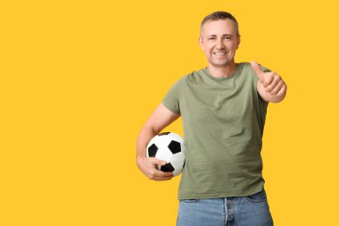 Futbol topuyla yakışıklı, olgun bir adam sarı arka planda baş parmak hareketi yapıyor.