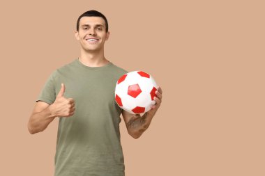 Elinde futbol topu olan yakışıklı genç adam kahverengi arka planda baş parmak hareketi yapıyor.